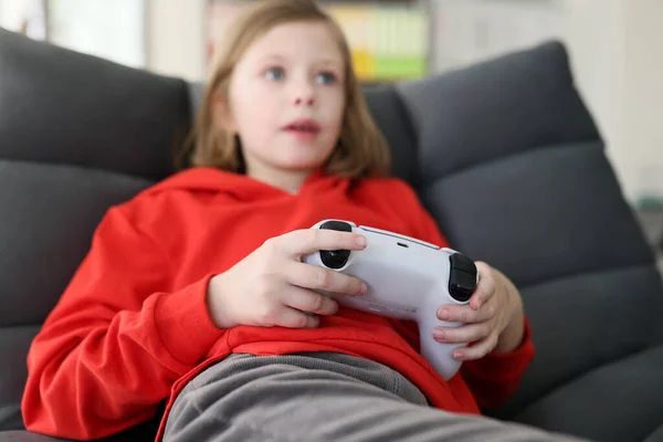 stock image Focused junior schoolgirl plays video game at home sitting in armchair. Little girl in red hoodie holds gaming console controlling process with open mouth