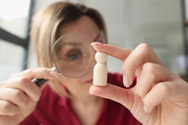 Focused woman examines small wooden figure with magnifier glass. Blonde female person holds white human figurine in hand looking through loupe