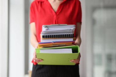 Kırmızı bluzlu sekreter elinde arşiv belgeleriyle klasörler tutuyor. Kadın muhasebe bilgileriyle evrak işlerini hallediyor.