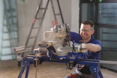 Gözlüklü mutlu ustabaşı testereyle tahta testereyi testereyle keserken gülümsüyor. Üniformalı pozitif adam odayı döşemeye hazırlanıyor.