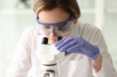 Koruyucu gözlüklü kadın bilim adamı elektronik mikroskop kullanarak örnek inceler. Laboratuvar kadın çalışanı bilimsel araştırma yapıyor.
