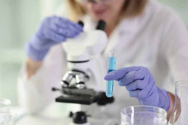 焦点を当てた女性は 青色液体を手にガラス管を保持する電子顕微鏡下でサンプルを調べる 研究室で研究を行う — ストック写真