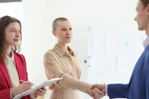 Happy manager watches colleagues shaking hands in office. Young woman with bald haircut and man feel happy to cooperate making profitable business deal