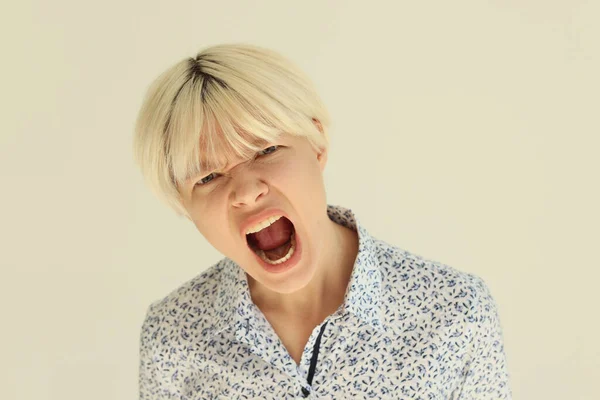 Emotional lady with dyed blonde hair screams with wide open mouth. Young woman shows acting skills on camera in studio on beige background