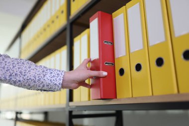 Ofis arşivinden muhasebe belgeleriyle birlikte kırmızı yüzük klasörünü alan bir kadın. Rafta malzemeler olan organize dizin yapısı