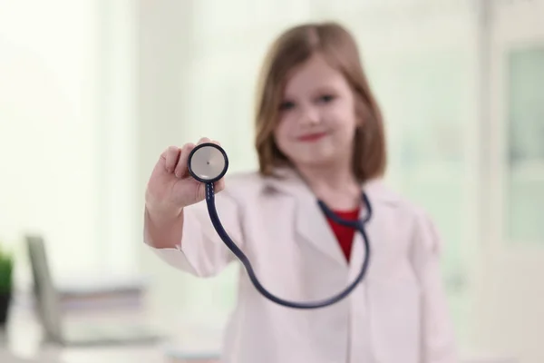 穿着白色实验室外套的可敬女孩 扮演医生 手持听诊器站在诊所的办公室 拿着医疗器械的小孩笑着看着镜头 — 图库照片