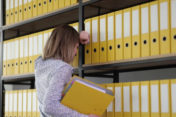 Exhausted woman fails finding right materials from archive for professional research. Secretary leans on rack with yellow ring binders on shelves