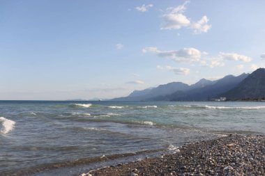 Dalgalı deniz ve beyaz köpüklü çakıl taşlı plaja yakın çekim, arka planda dağ manzarası. Resimli deniz kıyısı. Yaz zamanı, tatil konsepti.