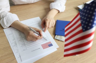 Genç bir kadın vize başvurusu formu dolduruyor. Tahta masada Amerikan bayrağı ve dolar banknotlu pasaportlu kadın tabelaları.