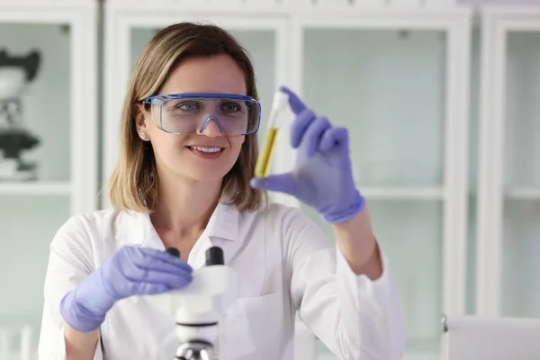 保護ガラスの笑みを浮かべて実験室のアシスタントは 試験管内の黄色の液体のサンプルを検査します 若い科学者は研究室で顕微鏡を使って — ストック写真