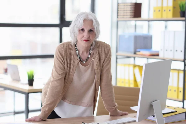 Serious elderly businesswoman stands leaning on wooden table with computer monitor. Office worker posing for photo in company premise
