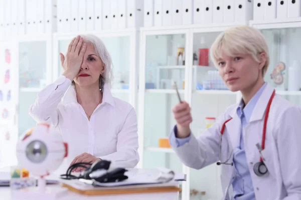 ブロンドの医者は職場のスネレン チャートに女性患者の手紙を示している 年配の女性は目を閉じて診療所に座っている間 — ストック写真