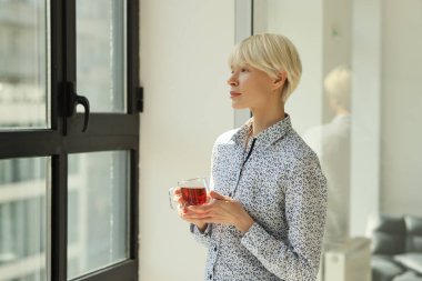 Çay fincanı taşıyan gizemli kadın modern şirket ofisinde pencereden dışarı bakıyor. Ciddi bayan müdürün dinlenme odasında dinlenme izni var.