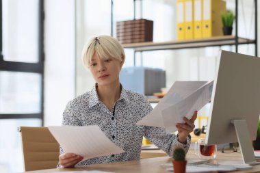 Ciddi sarışın kadın, kağıt çarşaflarda muhasebe belgelerini okuyarak bilgi karşılaştırıyor. Odaklanmış kadın çalışan bilgisayar monitörünün yanındaki masada oturuyor ve evrak işleriyle uğraşıyor.