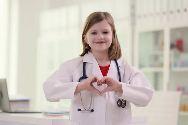 穿着白衣微笑的初中生扮演医生的角色 用手指表现出心征 带听诊器的积极女孩站在现代办公室里 — 图库照片