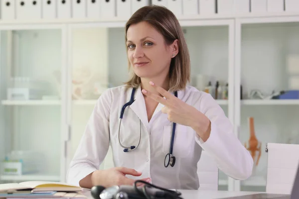 正の女性医師は カメラと笑顔で見て2本の指勝利ジェスチャーを示しています 専門医が診療所のテーブルに座る — ストック写真