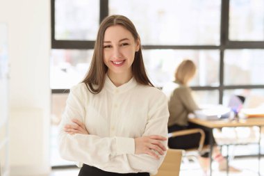 Neşeli bayan yönetici kollarını kavuşturarak modern ofiste duruyor. Beyaz bluzlu olumlu genç bir kadın kameraya bakar yüzünde gülümsemeyle.