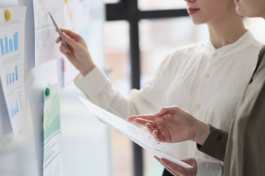 Meslektaşım verileri incelerken, kadın kalemle beyaz tahtadaki araştırma kağıdını işaret ediyor. Çalışanlar modern ofiste iş seminerine hazırlanıyorlar.