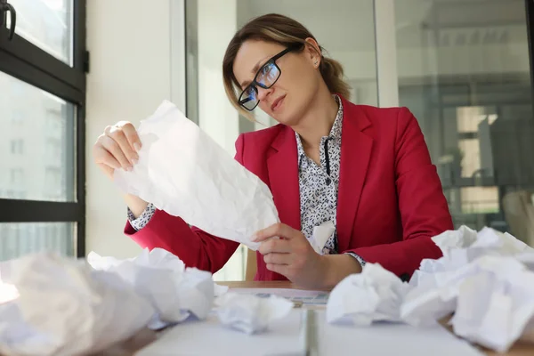 Disappointed female manager in glasses reads sheet sitting at table littered with scattered crumpled papers. Woman fails writing good ideas for project