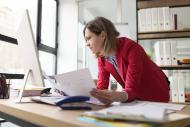 İstatistikler ve çizelgeler ile muhasebe kağıtlarını kontrol eden dikkatli bir bayan yönetici. Pembe ceketli kadın ofisteki bilgisayarın yanındaki masaya yaslanıyor.