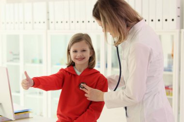 Mutlu küçük kız klinikteki sağlık kontrolünde baş parmağını kaldırıyor. Bakıcı kadın aile doktoru steteskopla hastanın kalbini kontrol ediyor.