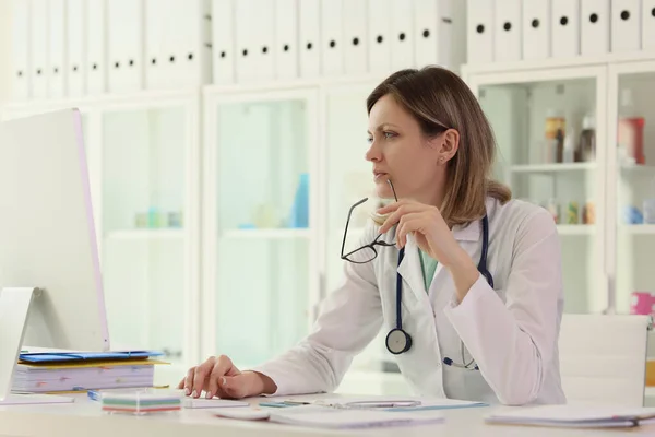 集中的な女性医師は 診療所のコンピュータに関する患者情報をチェックします 制服姿の女がテーブルに座る手に眼鏡を持って — ストック写真