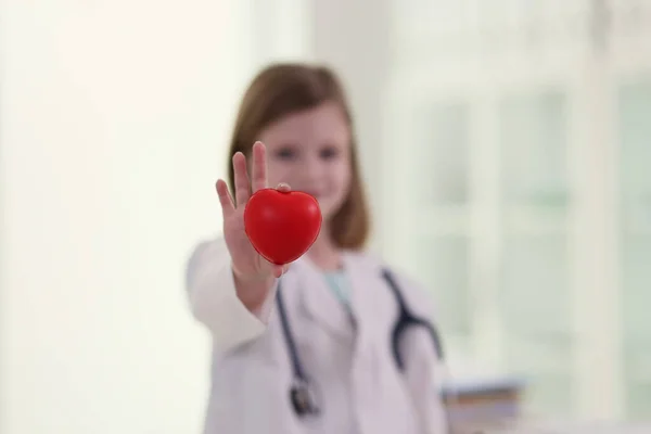 漂亮的小女孩穿着医生的衣服 手里拿着红心玩具 带听诊器的儿童站在诊所的办公室里 心脏病的治疗 — 图库照片