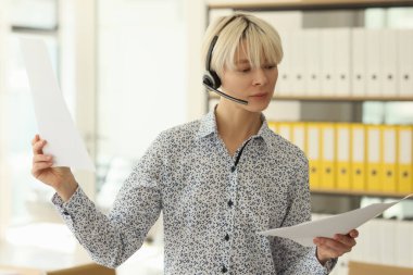 Kablosuz kulaklık takan odaklanmış yönetici müşterileri dinler ve proje belgelerini okur. Sarı saçlı genç kadın çağrı merkezinde çalışıyor.