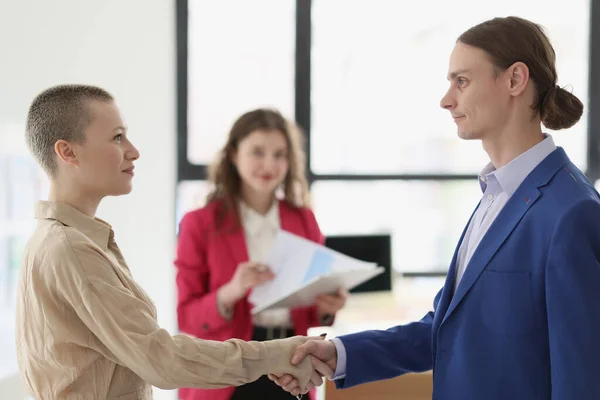 Serious partners man and woman shake hands making successful business deal while woman checking papers. Happy colleagues smile standing in modern office
