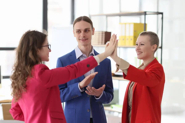 Female colleagues give high five while man boss applauds. Office employees rejoice at good deal standing at workplace in spacious premise