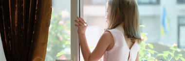 Close-up of little girl in pink dress standing on windowsill at open window. Danger of falling from height concept