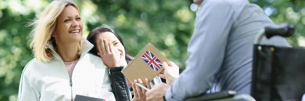 Portrait of disabled man in wheelchair studying english with friends in park. Happy male holding notepad with england flag. Disability and healthcare concept