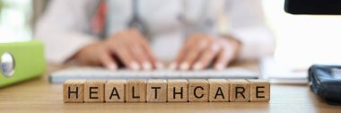 Close-up of healthcare word collected with wooden cubes in row. Woman doctor in medical gown on background. Medicine, treatment and wellbeing concept clipart