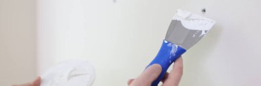 Hands with a spatula and putty against the background of a white wall, a close-up. Scatter holes on the wall