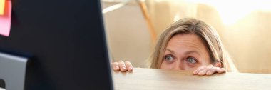 Woman hides behind office desk and looks at computer monitor. Scary movie, mistake at work, dodging online conversations concept.