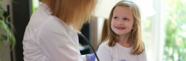Young female patient smiles at doctor at medical examination. Nurse listens to heartbeat of happy girl in medical clinic.