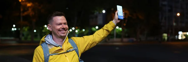青いバックパックと携帯電話を手に黄色のコートの男は夜に路上で彼の友人に会う タクシーサービスのためのモバイルアプリケーションを使用して 観光客がタクシーを呼び出し — ストック写真