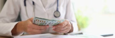 Doctor counts money in her hands in medical clinic. Close-up of many dollar bills in the hands of nurse. Cost of medicine and health insurance concept.