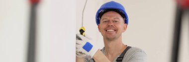 Happy painter in hard hat paints wall with white paint and looks at camera. Decorator stands with roller brush and smiles.