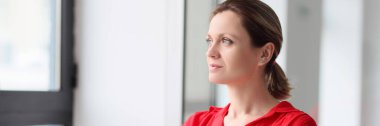 A young thoughtful woman in red clothes looks out the window, a close-up. Concept of thinking