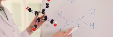 Female chemist holding molecular model and drawing chemical formulas on whiteboard. Chemistry, education and science research concept.