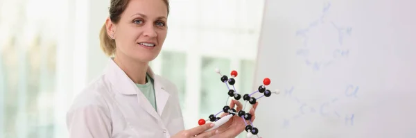 Female scientist standing near whiteboard, pointing to molecular model in her hand and looking at camera. Scientific research and education concept.