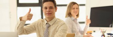 Two managers are sitting at their desks in office and showing thumbs up gesture. Concept of successful business and career.