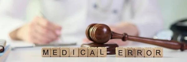 Words medical errors collected from wooden cubes with judges gavel on table, doctor with stethoscope in background. Malpractice, medical error and investigation concept.