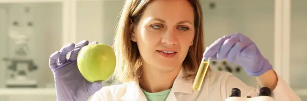 戴橡胶手套的女人手里拿着带黄色液体样品的新鲜苹果和软管 实验室的专家站在显微镜下 — 图库照片