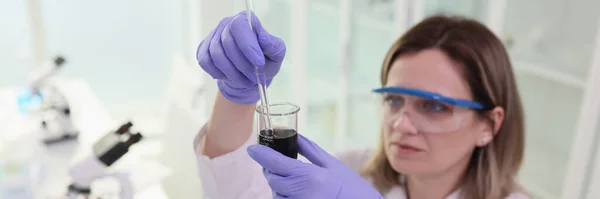 Focused female scientist in goggles mixes black liquid in glassware. Woman in rubber gloves does scientific research of crude oil in lab