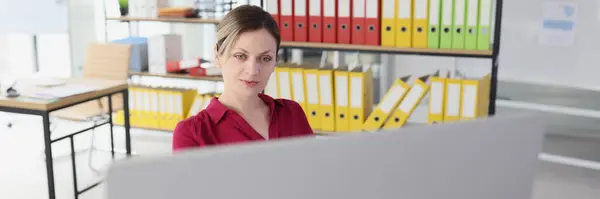 Blonde woman sits at table hidden behind big computer monitor in office against rack with ring binders. Female secretary at workplace in office