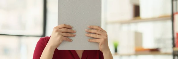 Female secretary hides face behind clipboard sitting at table in office. Woman in red blouse feels exhausted at work waiting for day to finish for rest