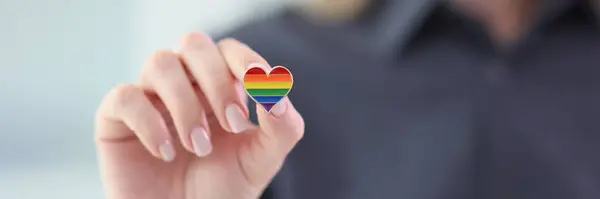 Woman holds small heart badge with LGBT rainbow flag colors in hand. Female person shows respect and support for community representatives