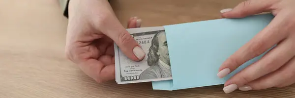 Woman with neat manicure takes dollar banknotes out of envelope sitting at wooden table in office. Female employee checks bribe income from deal
